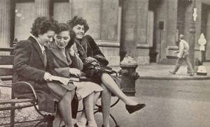 Three women sitting on a bench. They are wearing coats and two are looking at something together.