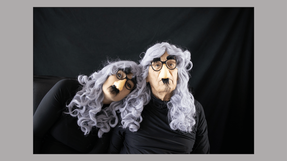 A young woman leans on an elder man. Both of them are wearing the same all black turtlenecks, grey curly wigs with bangs and groucho marx glasses.