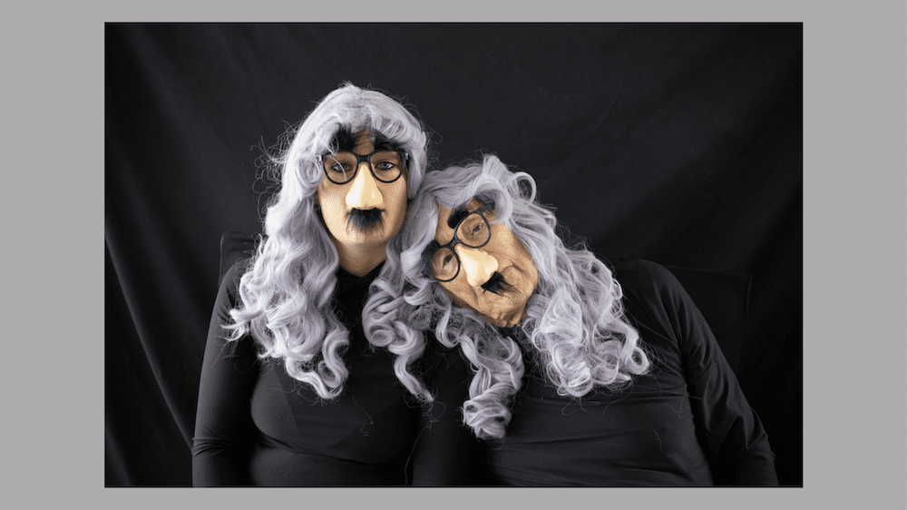 An elderly man leans on a young woman. Both of them are wearing the same all black turtlenecks, grey curly wigs with bangs and groucho marx glasses.