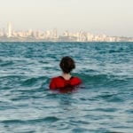 Woman standing shoulder level in the ocean facing away from the camera with a city in the background