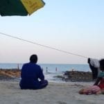 Three people facing away from the camera sitting on sand near the ocean