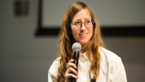 A long haired woman, Alison O'Daniel, smiles while holding a microphone. She is wearing glasses and a white blouse.