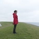 Woman in red coat stands on grass staring into the distance