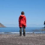 Woman in red coat stands towards the ocean facing away from the camera