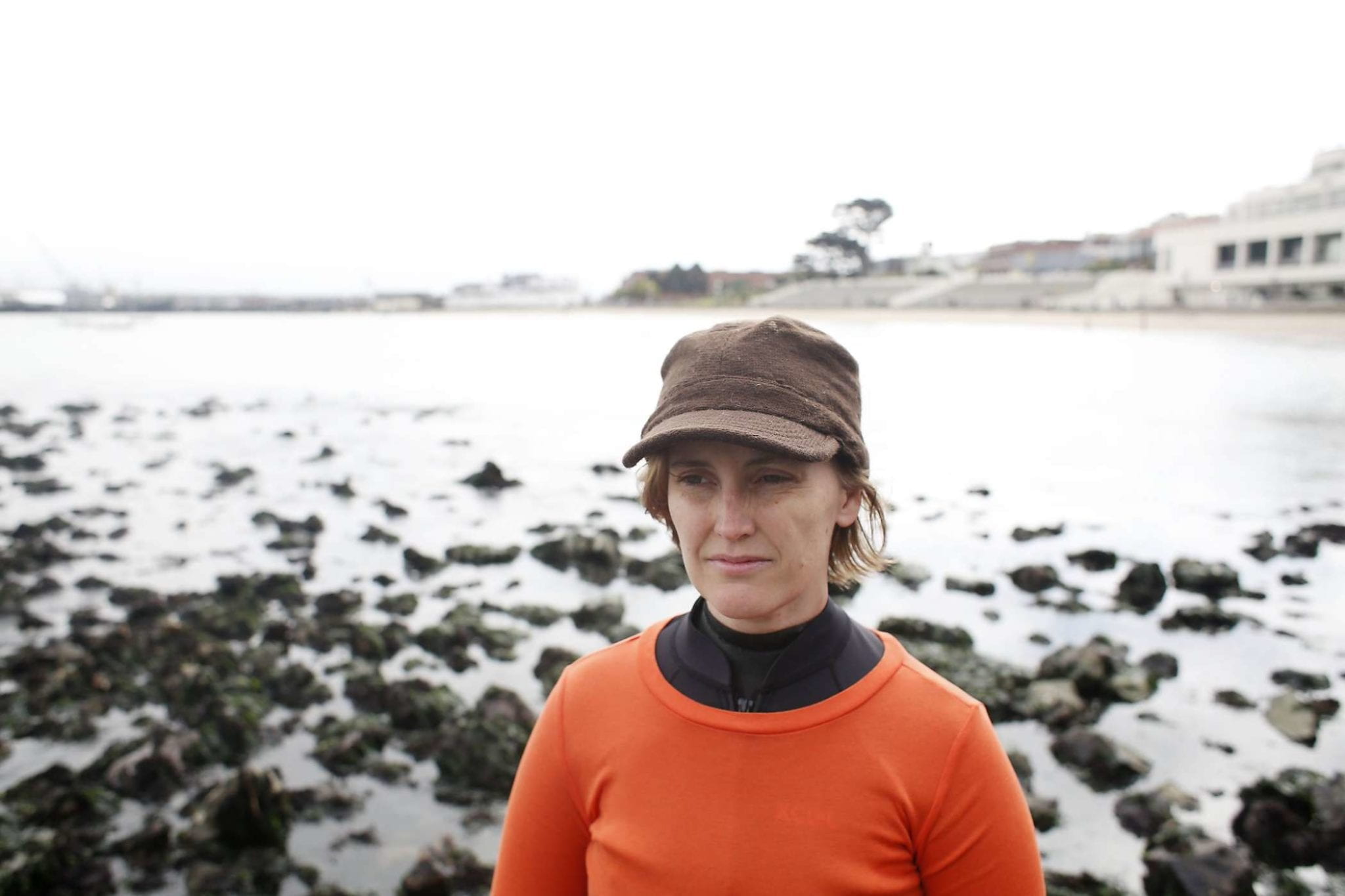 Sarah Cameron Sundae stands in a rocky ocean. She is wearing a brown hat and a orange top over her wetsuit.