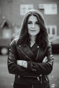 Katia Repina is wearing a leather jacket. Her arms are crossed and is looking into the camera. Out of focus are cars and a house. 