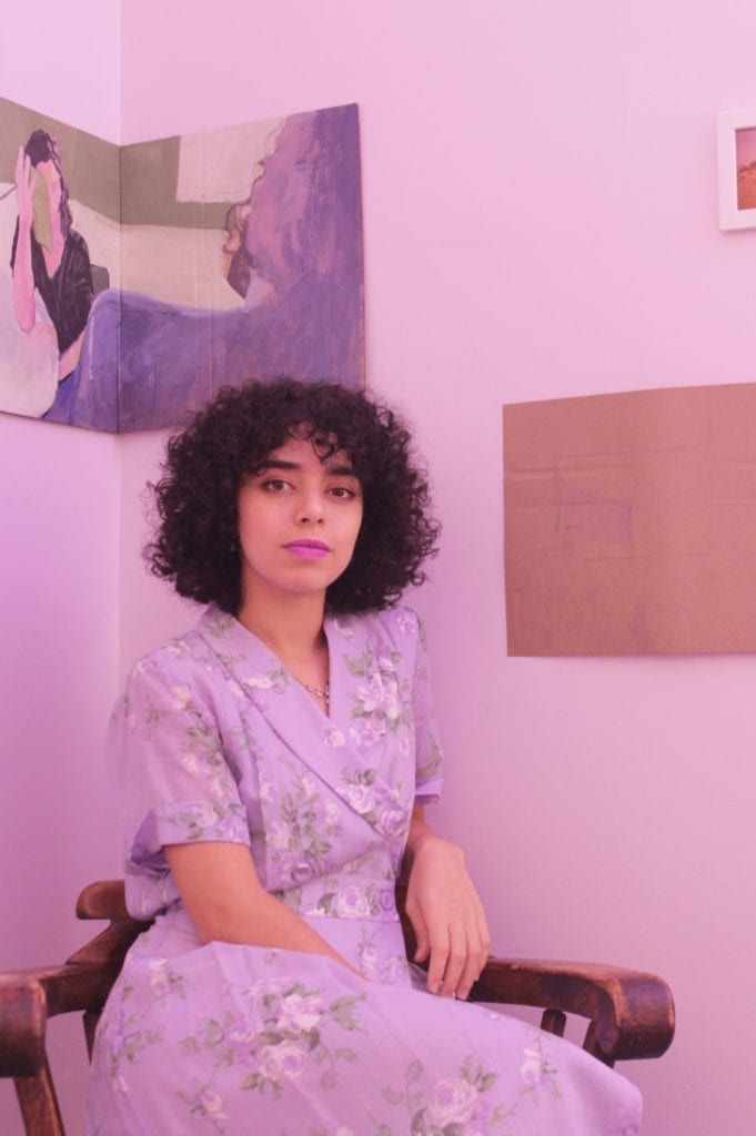 A dark short curly hair woman in a purple floral dress sits in a room corner.