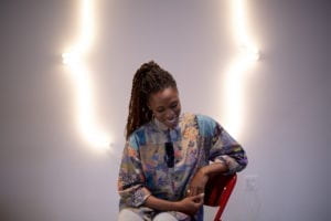 A woman sits on a chair with a neon sculpture behind her. She is smiling at the ground and is wearing a colorful shirt.