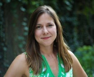 Anna Sale gives a slight smile to the camera. She is wearing a green top that compliments the foliage behind her