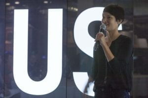 Chinese-American woman with short dark hair holding a microphone in front of large digital letters that read “Us”.