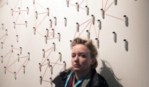 Blonde woman stands against a white wall with vials.