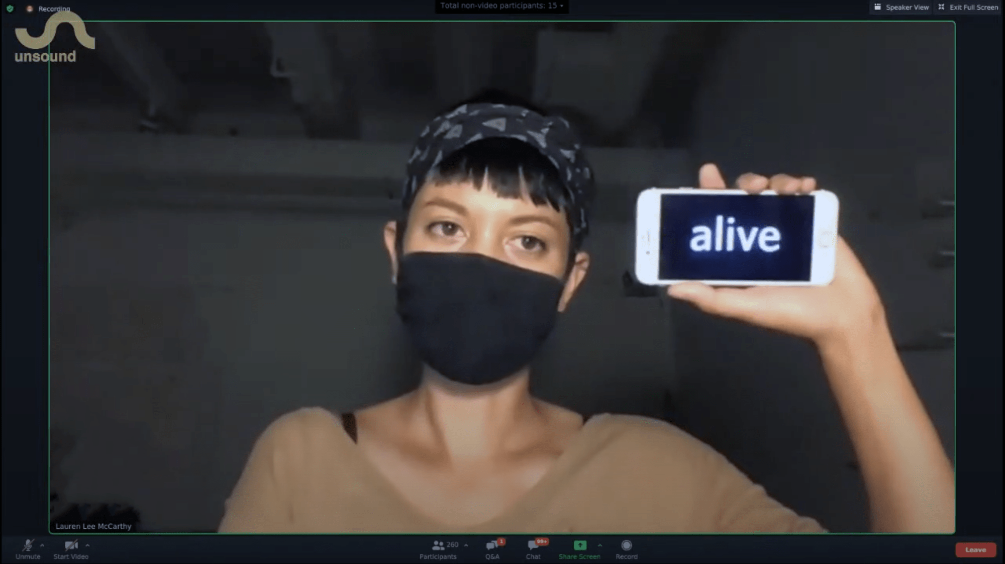 A woman with a black mask on holds up her smartphone that has the text "alive" on it