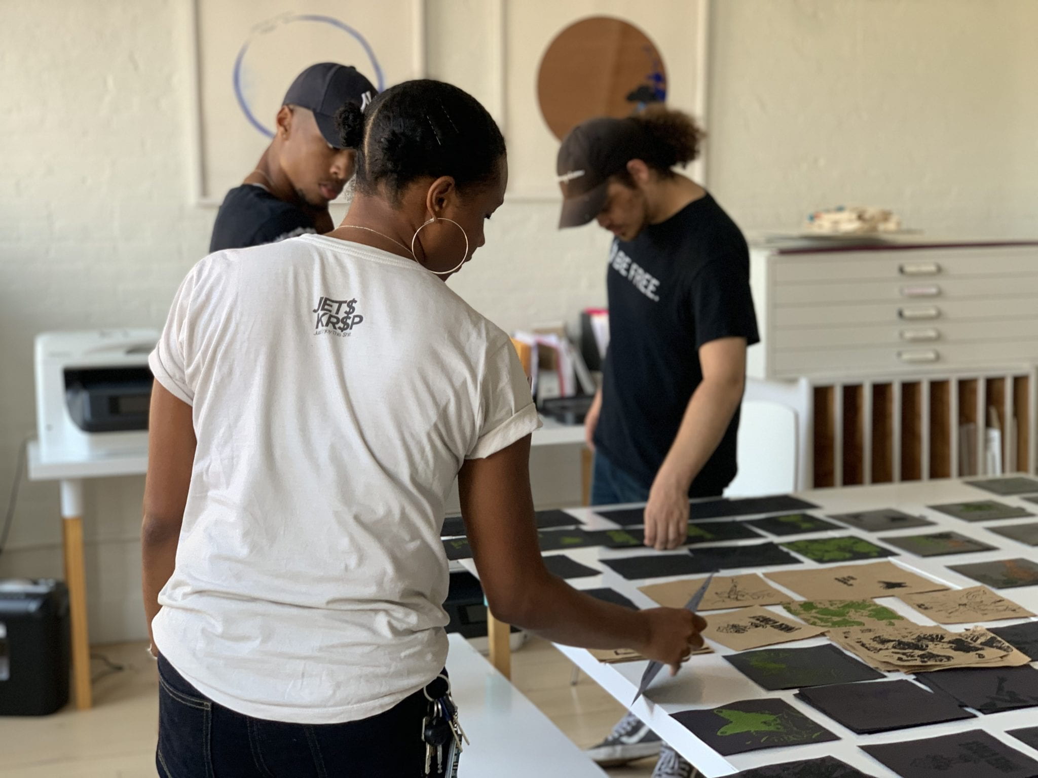 Three people look at small works of art on a table.