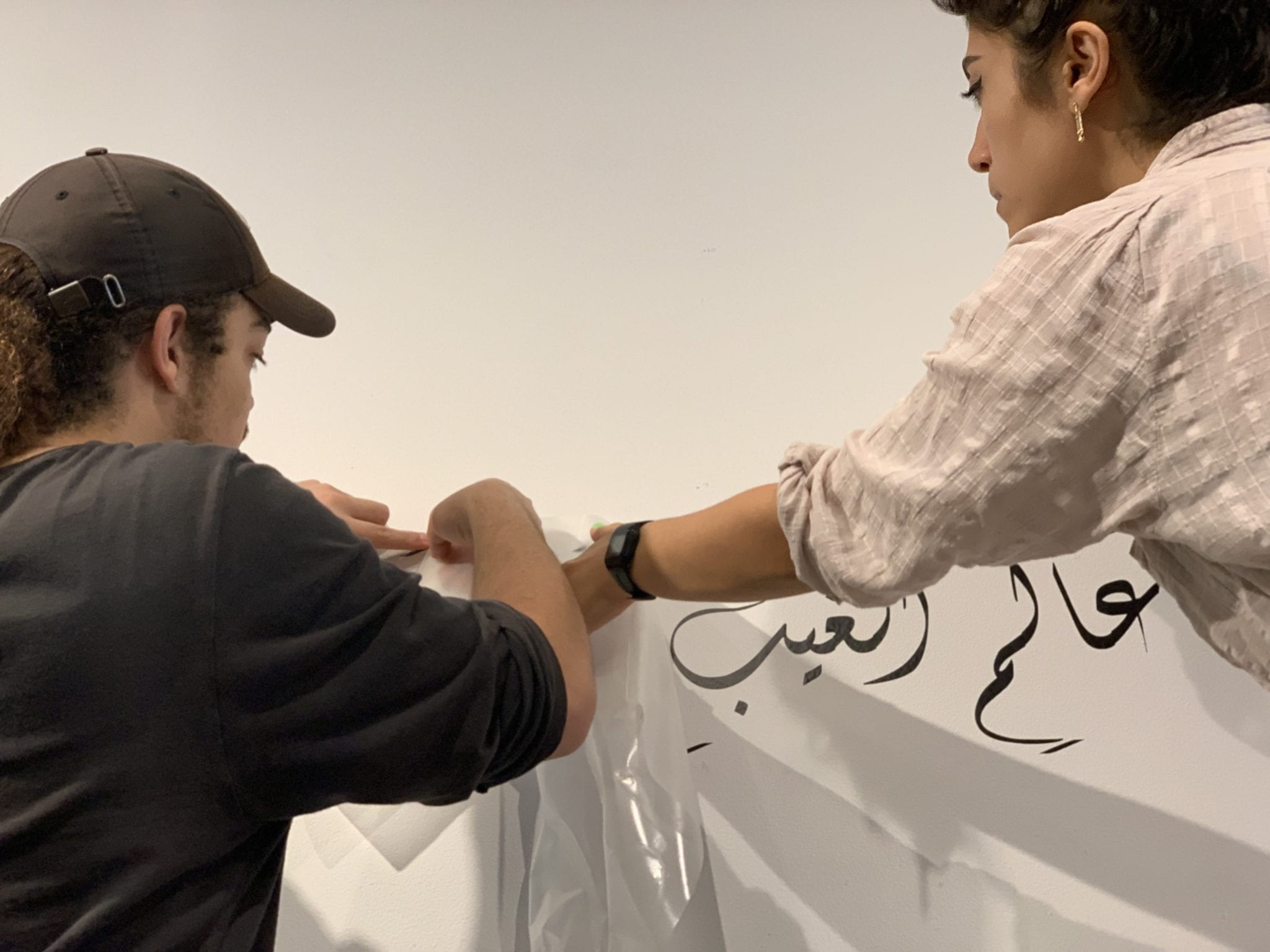 Two people from behind put plastic on a painting of Arabic letters.