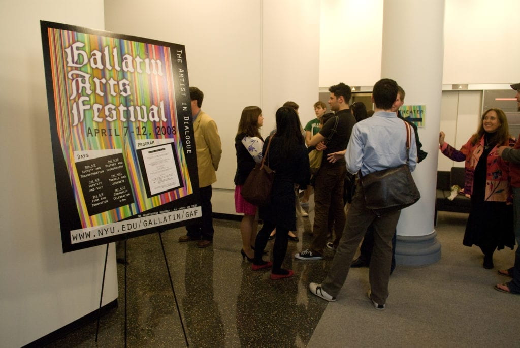 A group of people stand in a lobby are conversing next to an easel with a sign that reads "Gallatin Arts Festival"