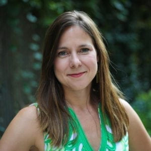 Anna Sale gives a slight smile to the camera. She is wearing a green top that compliments the foliage behind her