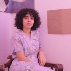 A dark short curly hair woman in a purple floral dress sits in a room corner.