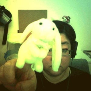 A woman wearing headphones holds up a bunny finger puppet to the camera