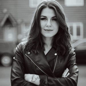 Katia Repina is wearing a leather jacket. Her arms are crossed and is looking into the camera. Out of focus are cars and a house.