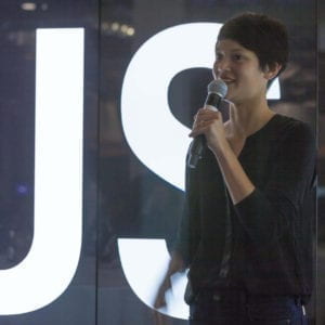 Chinese-American woman with short dark hair holding a microphone in front of large digital letters that read “Us”.