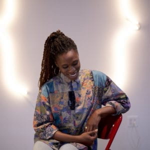 A woman sits on a chair with a neon sculpture behind her. She is smiling at the ground and is wearing a colorful shirt.