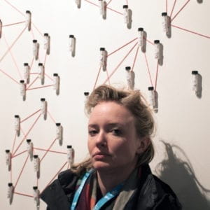 Blonde woman stands against a white wall with vials.