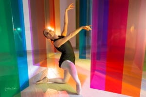 Dancer poses in front of multicolor plexiglass panels. She is wearing ballet shoes, unitard, and is in a ballet pose.