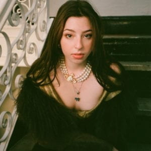 Photograph of the artist. She is wearing pearls and a fur jacket. She is sitting on a staircase with an elaborate iron railing