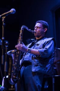 Emilio plays the saxophone on a stage. He is lit by blue light