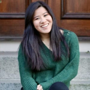 Photograph of the artist. She is wearing a green sweater and is sitting on steps in front of a wooden door