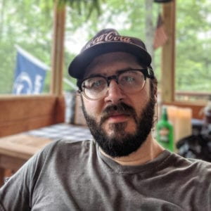 Selfie of a man with a beard, hat, and glasses giving a neutral face to the camera