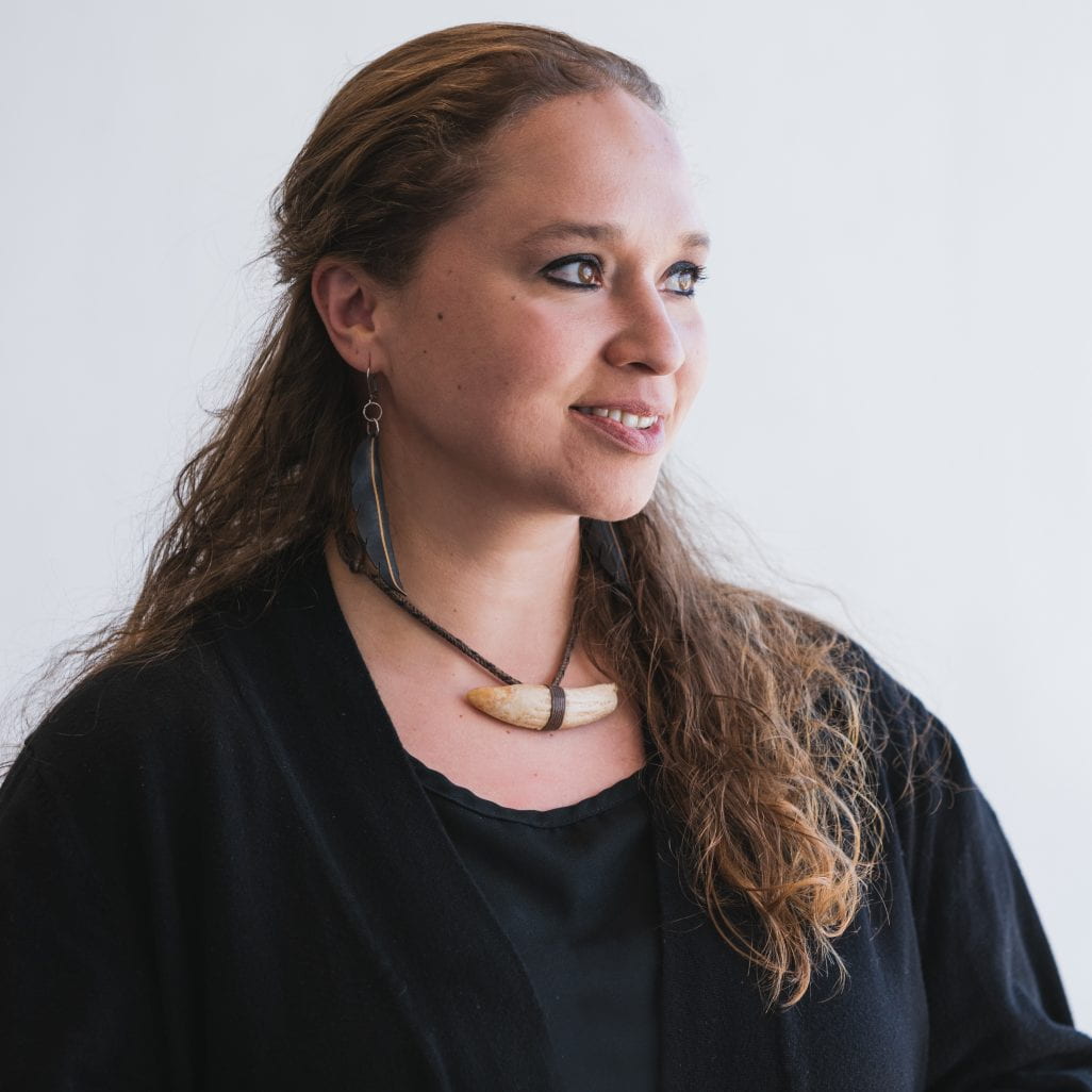 Photograph of woman with feather earrings and a bone necklace looking off into the distance