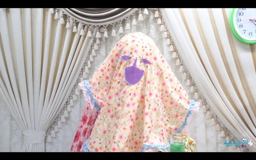 Still from "Um Al Naar (Mother of Fire)." A figure stands before a background of white decorated fabric. The figure is cloaked in several colorfully patterned fabrics.