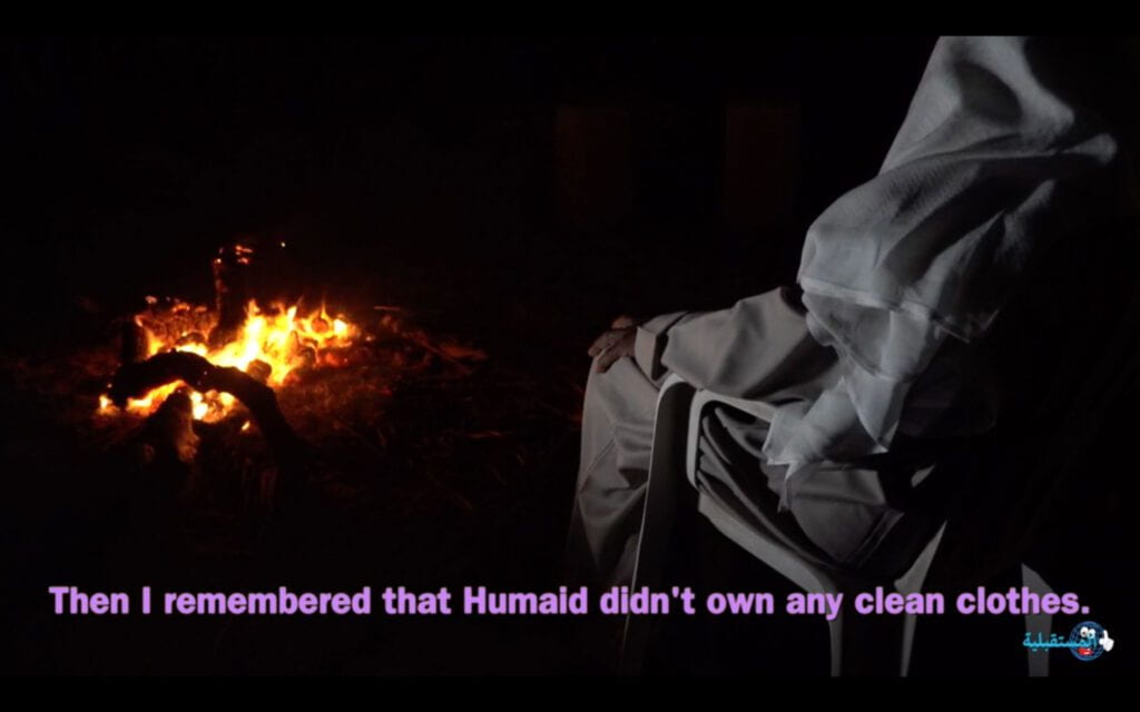 Still from "Um Al Naar (Mother of Fire)," A cloaked figure sits before a fire. Subtitles read, "Then I remembered that Humaid didn't own any clean clothes."