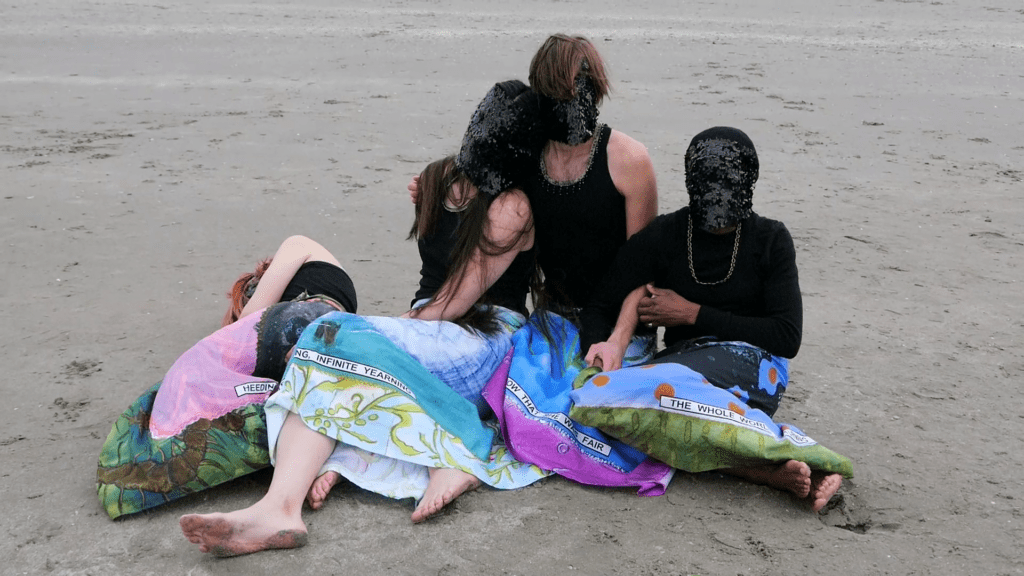 Three individuals stting on wet sand lean into one another. They all wear colorful skirts, black shirts, and black sequined masks that cover their entire faces.