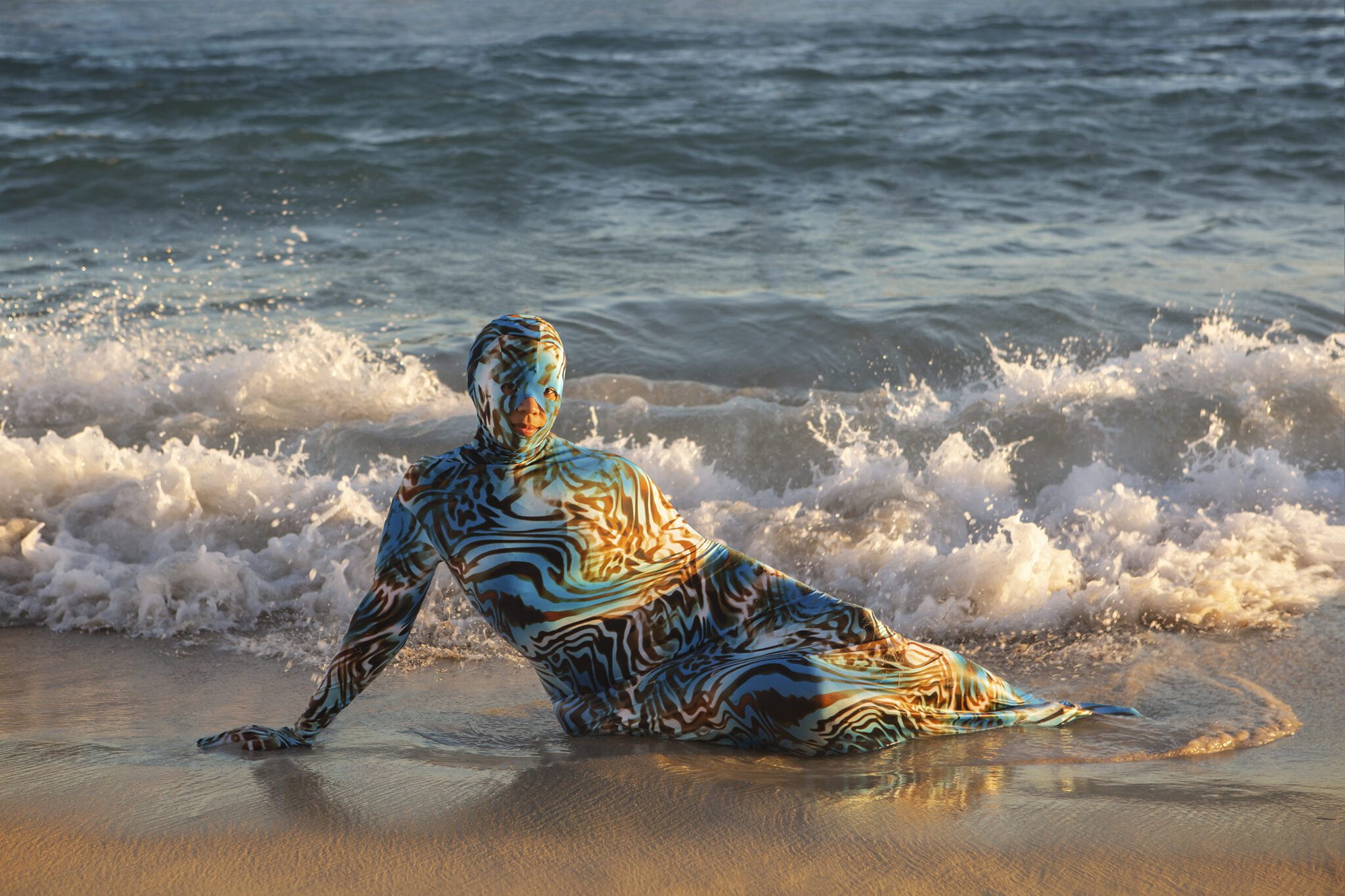 Human in a head-to-toe bodysuit, reclining on their side in front on the beach with waves crashing behind them.