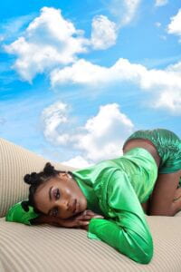 A figure in silky a green shirt and shorts lays on a couch. Behind them is stretches a blue sky with clouds