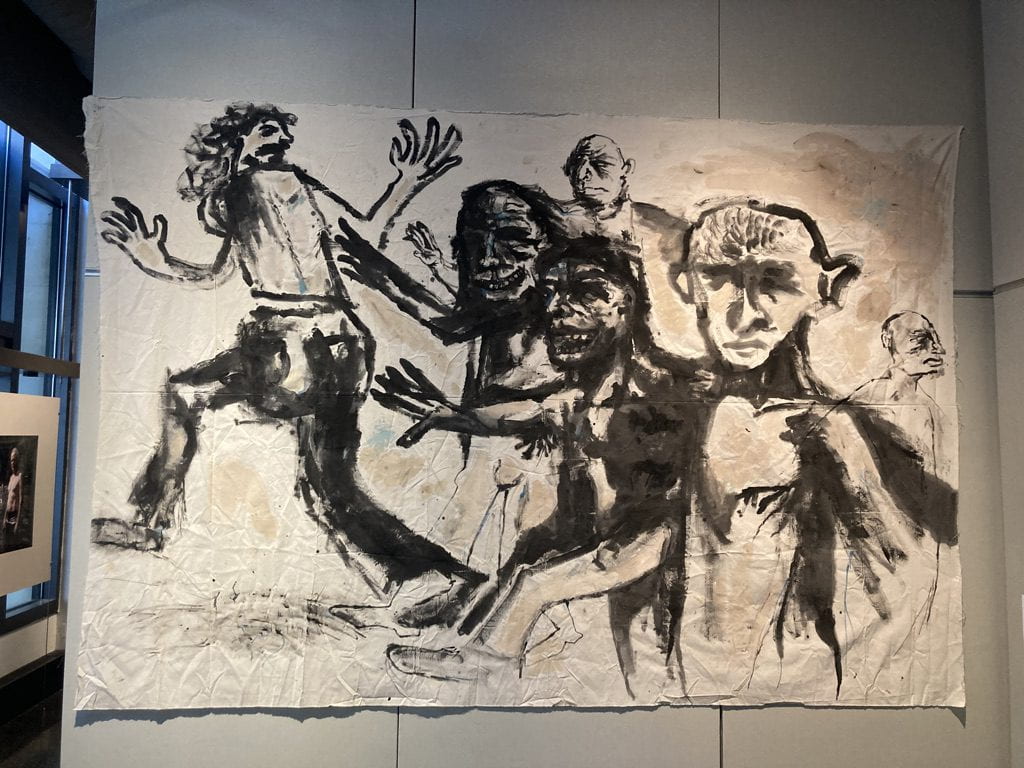 Large loose canvas hung on a white wall, containing five figures who are painted black chasing after each other. 