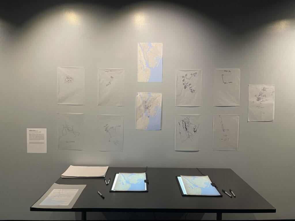 An installation photograph of a black table with printed maps of Manhattan placed on a light box, along with loose tracing paper. The traced images are hung on a white wall, above the table. 