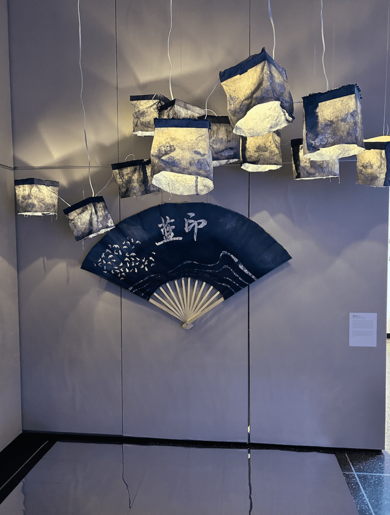 An installation image of a blue traditional Chinese fan with white lettering is hung in the center of a white wall. Blue and white lanterns float above the fan and a mirror is placed on the floor at the base of the wall. 