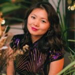 Chan Lin, an Asian American woman, sits in greenery with a button downed top with beads, smiling at the viewer.