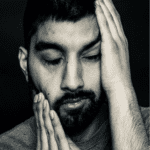 Black and white photo of a man with a beard and mustache, his eyes closed as he pushes his face up on the right and supports his chin on the right.