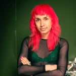 Photo of a person (she/her + they/them) with fucsia hair, arms crossed and a long sleeved black top with a green undergarment staring at the viewer on a green ground.