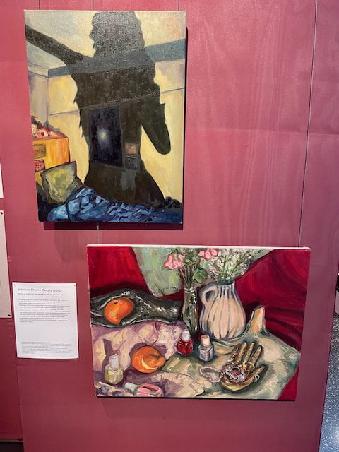 Photograph of two paintings hung on a maroon wall. One painting depicts the shadow of a figure looming over a room and the other painting depicts a flat surface with various items placed on top.