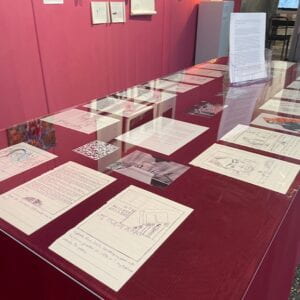 Photograph of a table lined with maroon fabric and plexiglass that lies on top of drawings on white paper and printed photographs. 