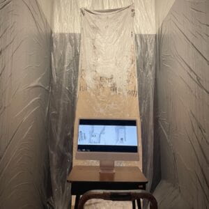 Photograph of a room installation, covered in plastic wrapping and a white sheet hanging on the center wall. There is a chair and desktop monitor on a desk at the center of the room. 