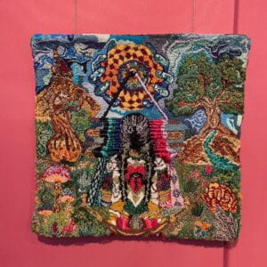 Photograph of an embroidered square on a maroon wall. The embroidery is multicolored and depicts a figure and a whimsical landscape. 