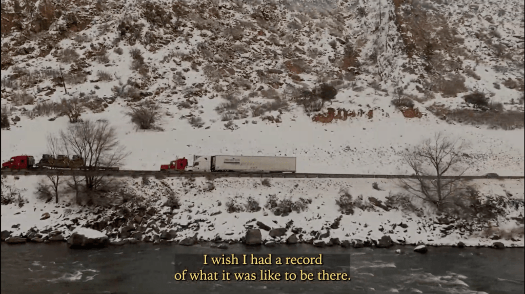 Photograph of trucks driving down a highway. The mountainous surrounding area is covered in snow and below the highway lies a flowing river. 