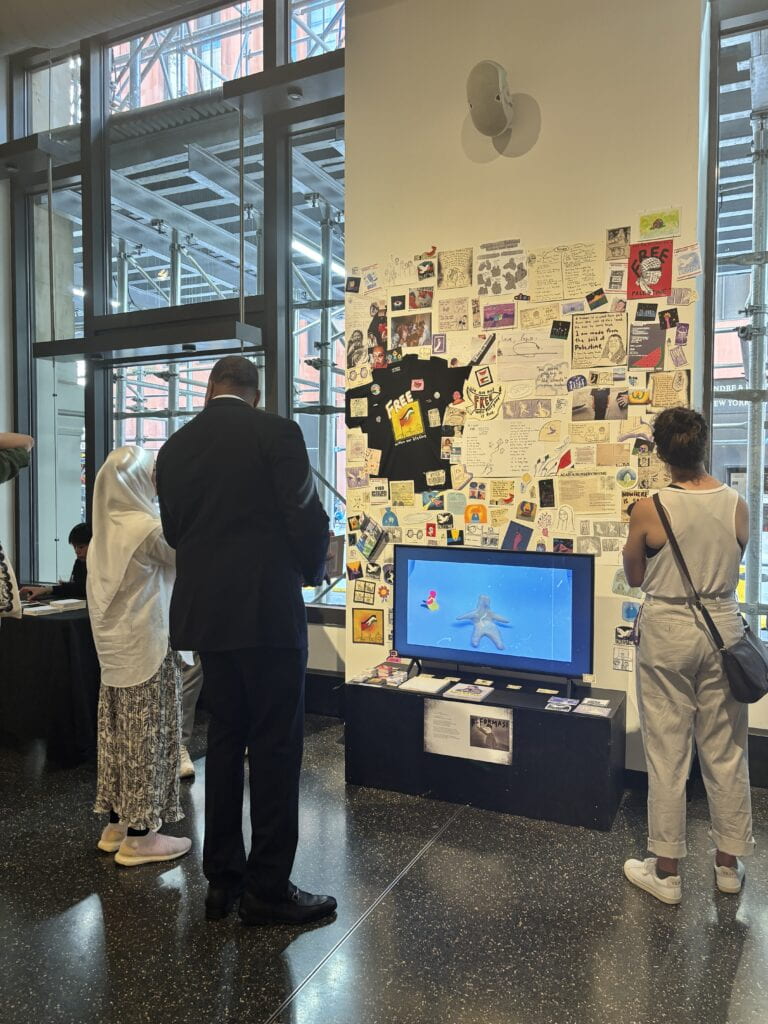 Image of opening night of gallery show depicting three people standing in front of a wall full of collaged images and a tv playing a video sitting on a black stand full of books and stickers.