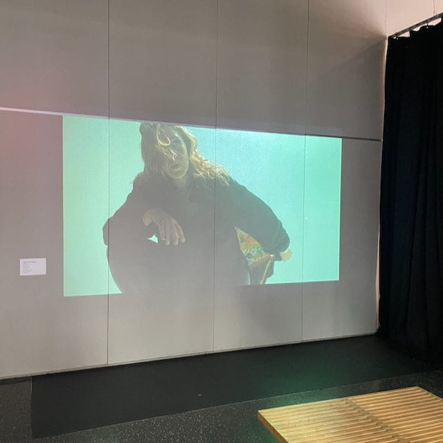  A digital image of a person with long hair displayed on a wall in a dimly lit room with a black curtain on the right.